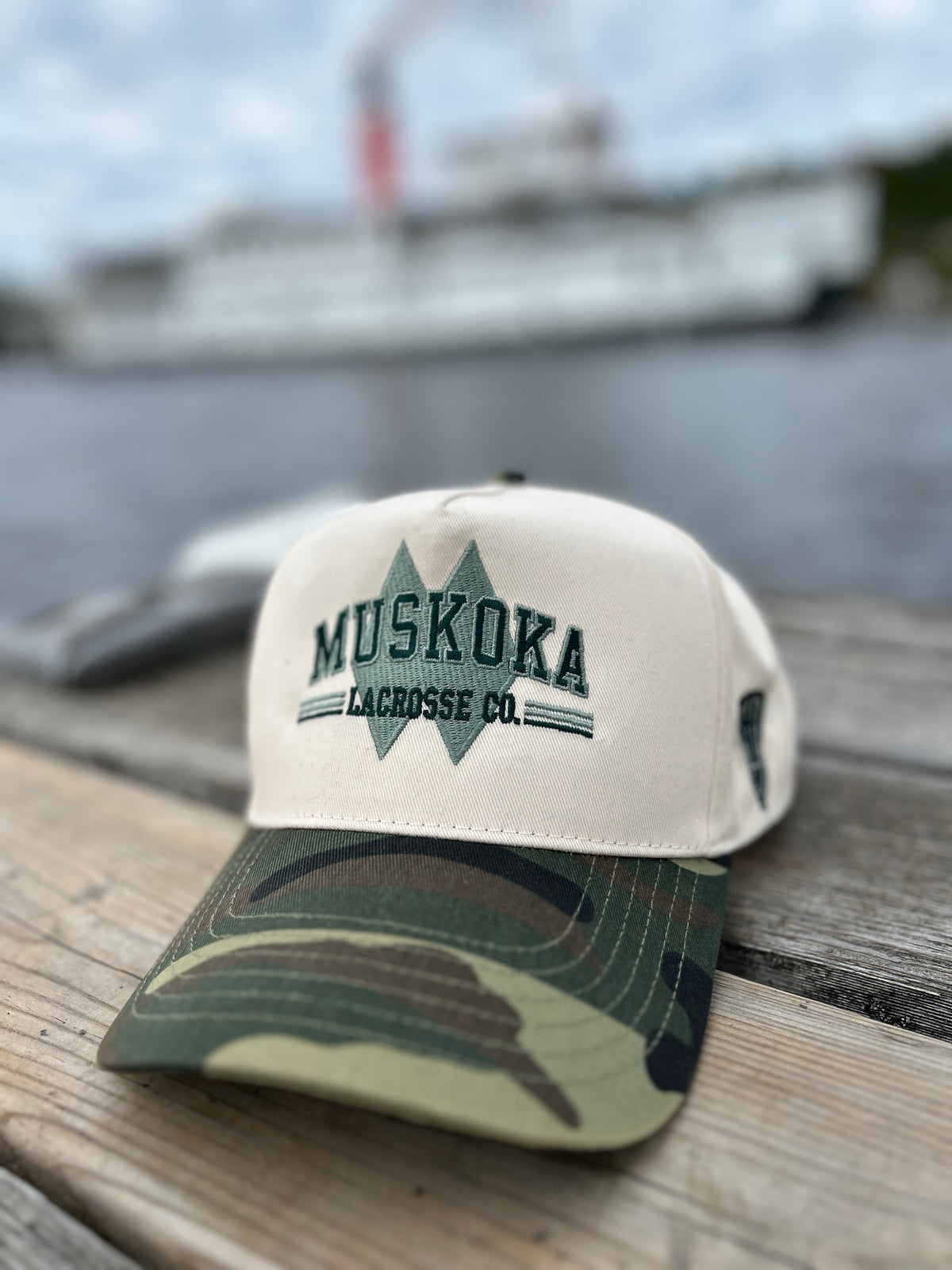 Muskoka Lacrosse Heritage Snapback with Ivory Crown, embroidered logo and camo brim. On a dock in Muskoka Steamboat 